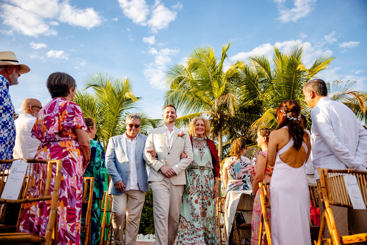 Couple walks down the aisle at an outdoor tropical wedding ceremony with palm trees and colorful guests.