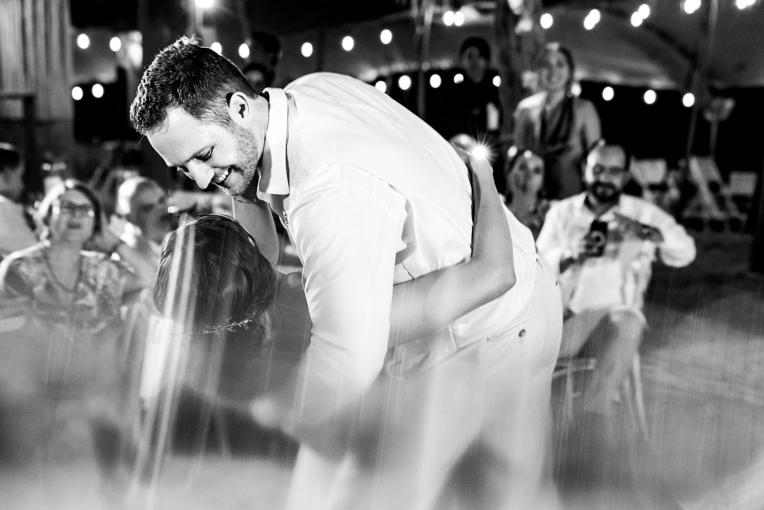 Black and white photo of a groom in white suit bowing at a wedding reception with guests in background.