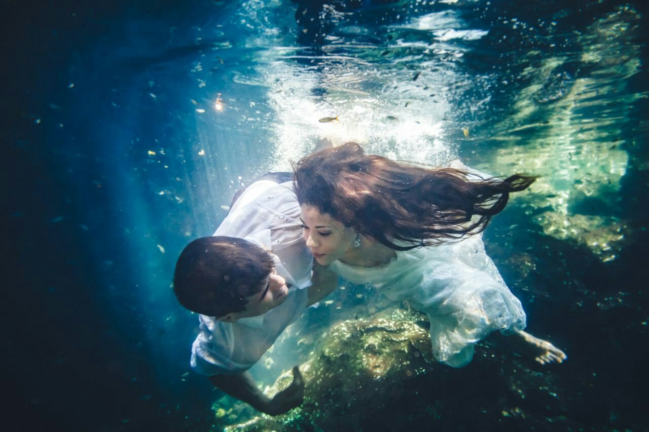 Couple sharing a romantic underwater kiss, woman in white dress with hair flowing, sunlight rays piercing blue water.