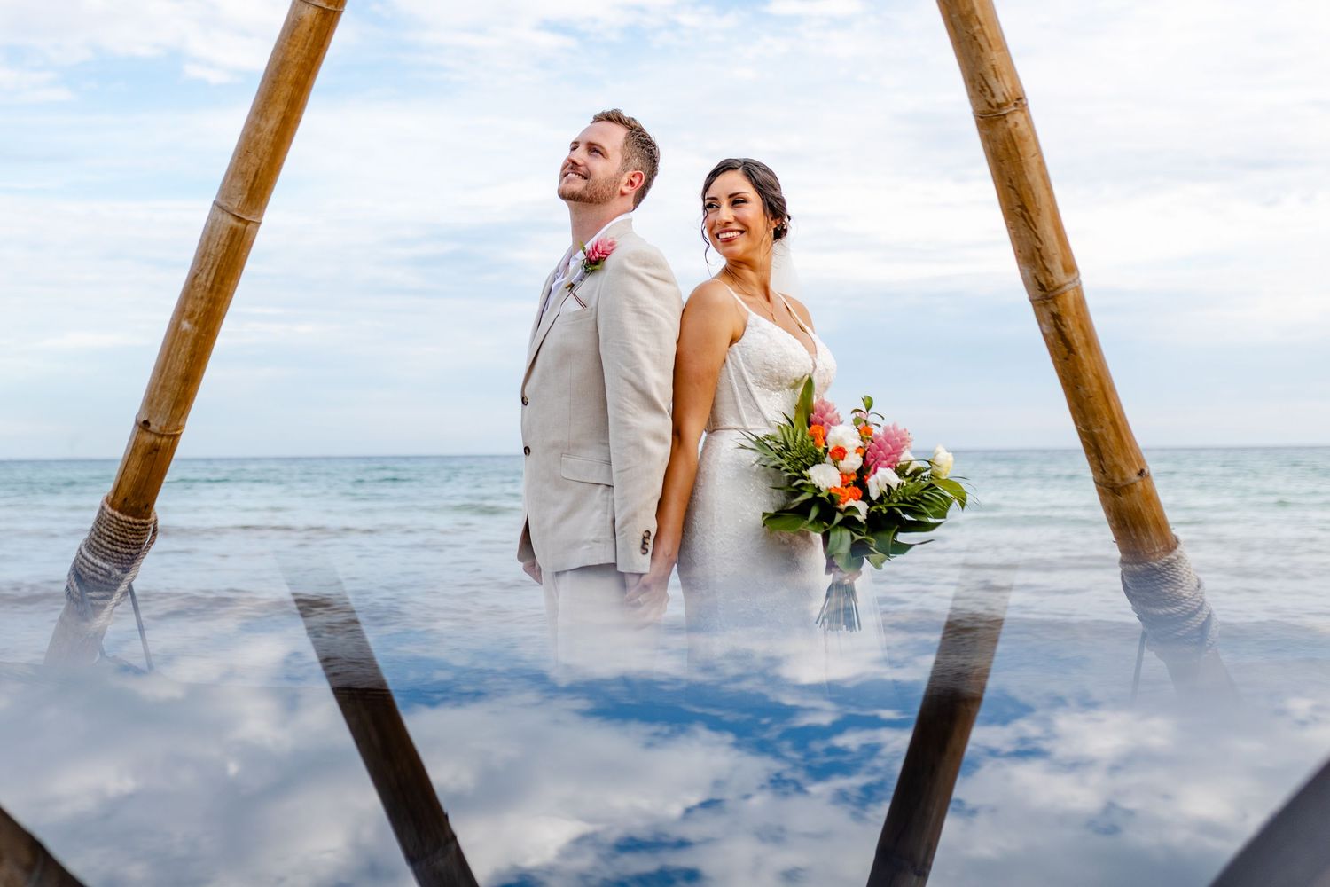 Bride and groom stand back-to-back under bamboo arch on tropical beach, bride holding colorful bouquet.