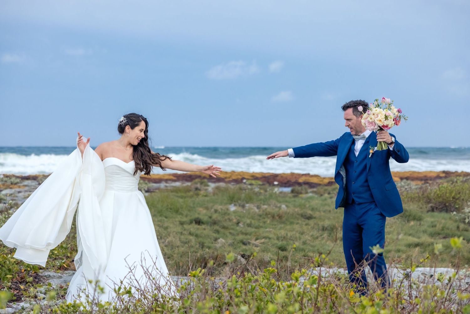 Bride and groom pose playfully on a windy beach, holding hands with ocean waves behind them on their wedding day.