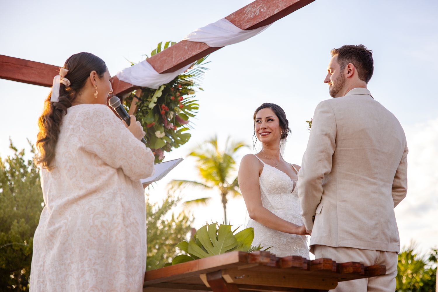 Bride and groom exchange vows at outdoor beach wedding ceremony under wooden arch with officiant.