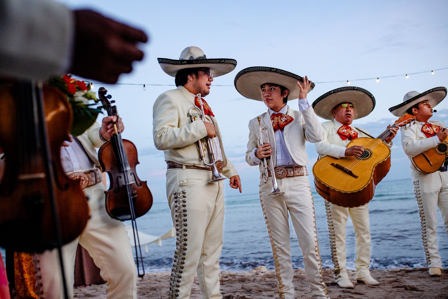 Mariachi musicians in white charro outfits playing violin and guitar on a scenic beachside venue.