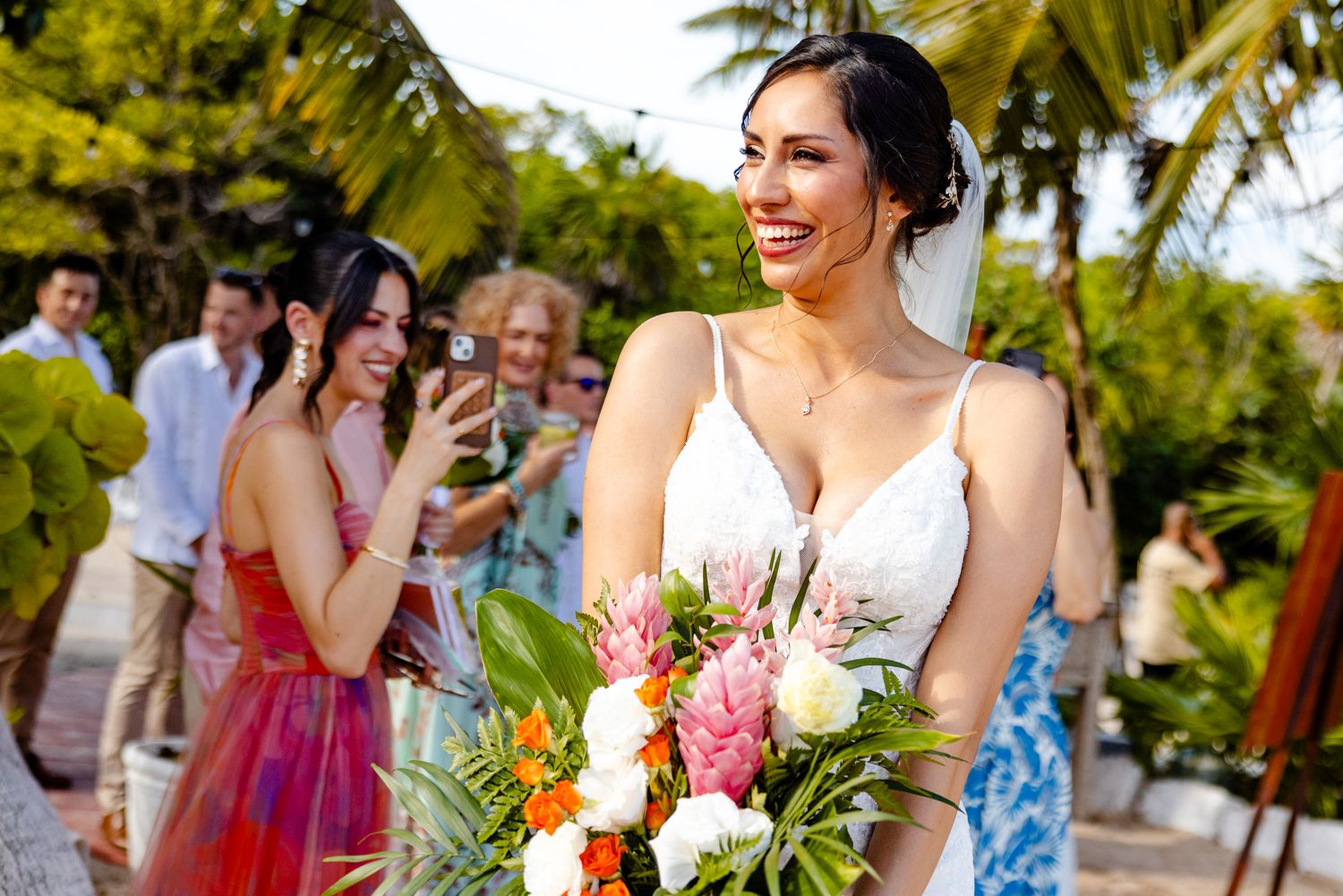 Smiling bride holding vibrant tropical bouquet with guests photographing her at an outdoor wedding ceremony.