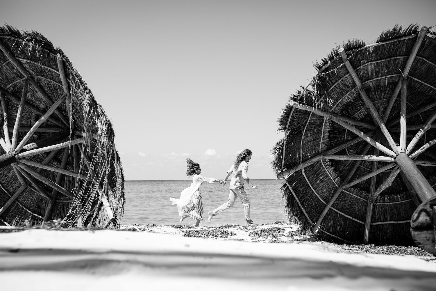 Two children run along a sandy beach between large round thatched huts by the ocean in black and white.