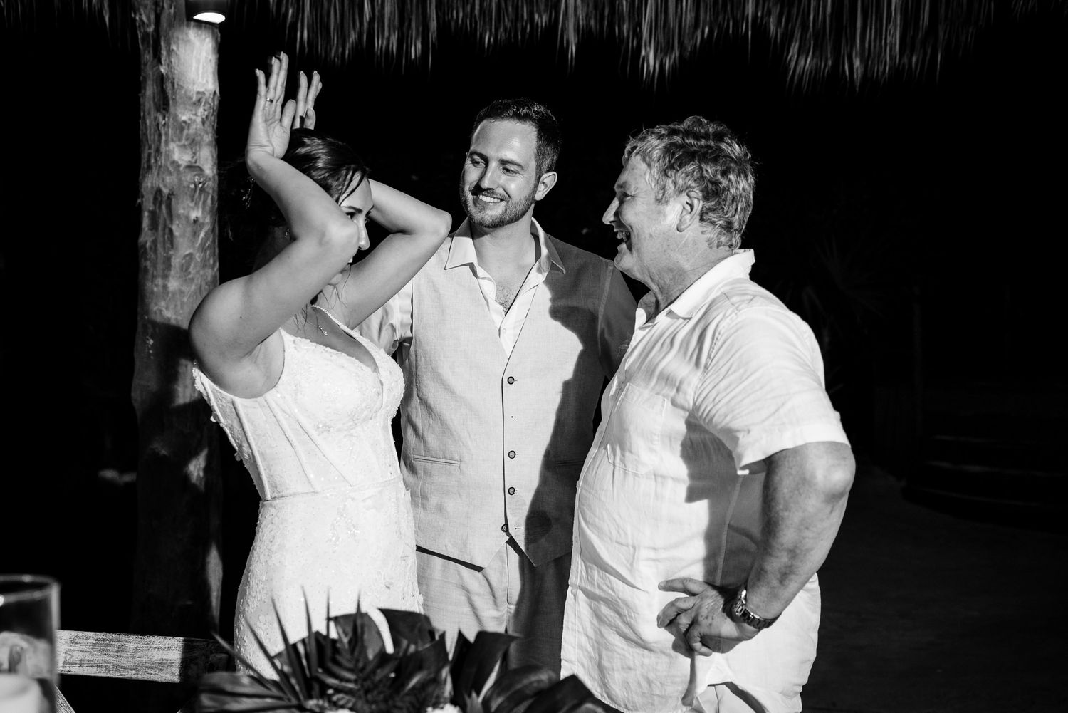 Black and white photo of bride and groom laughing with guest at outdoor tropical wedding reception.