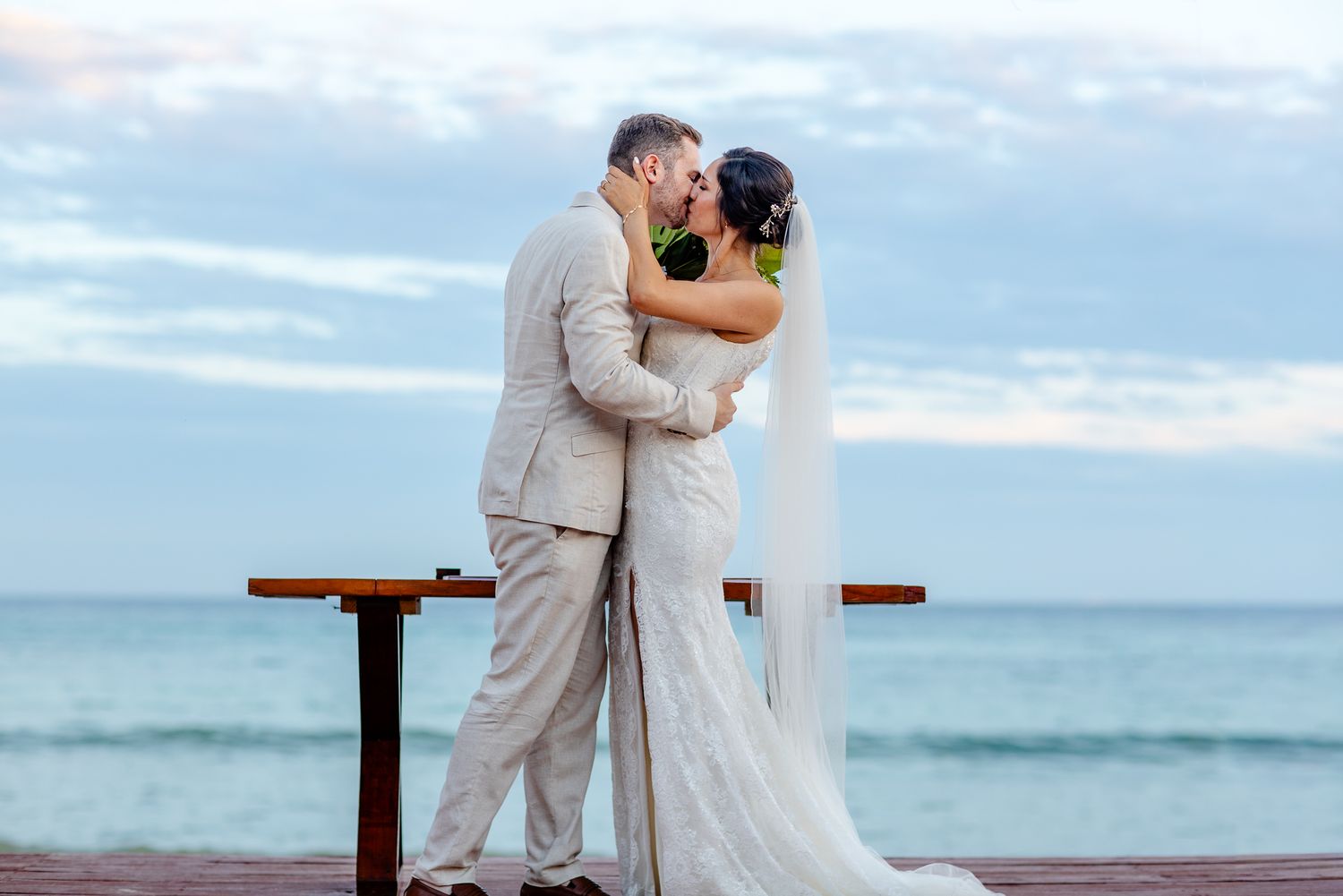 Bride and groom share a romantic kiss on a beach wedding ceremony with ocean backdrop at sunset.
