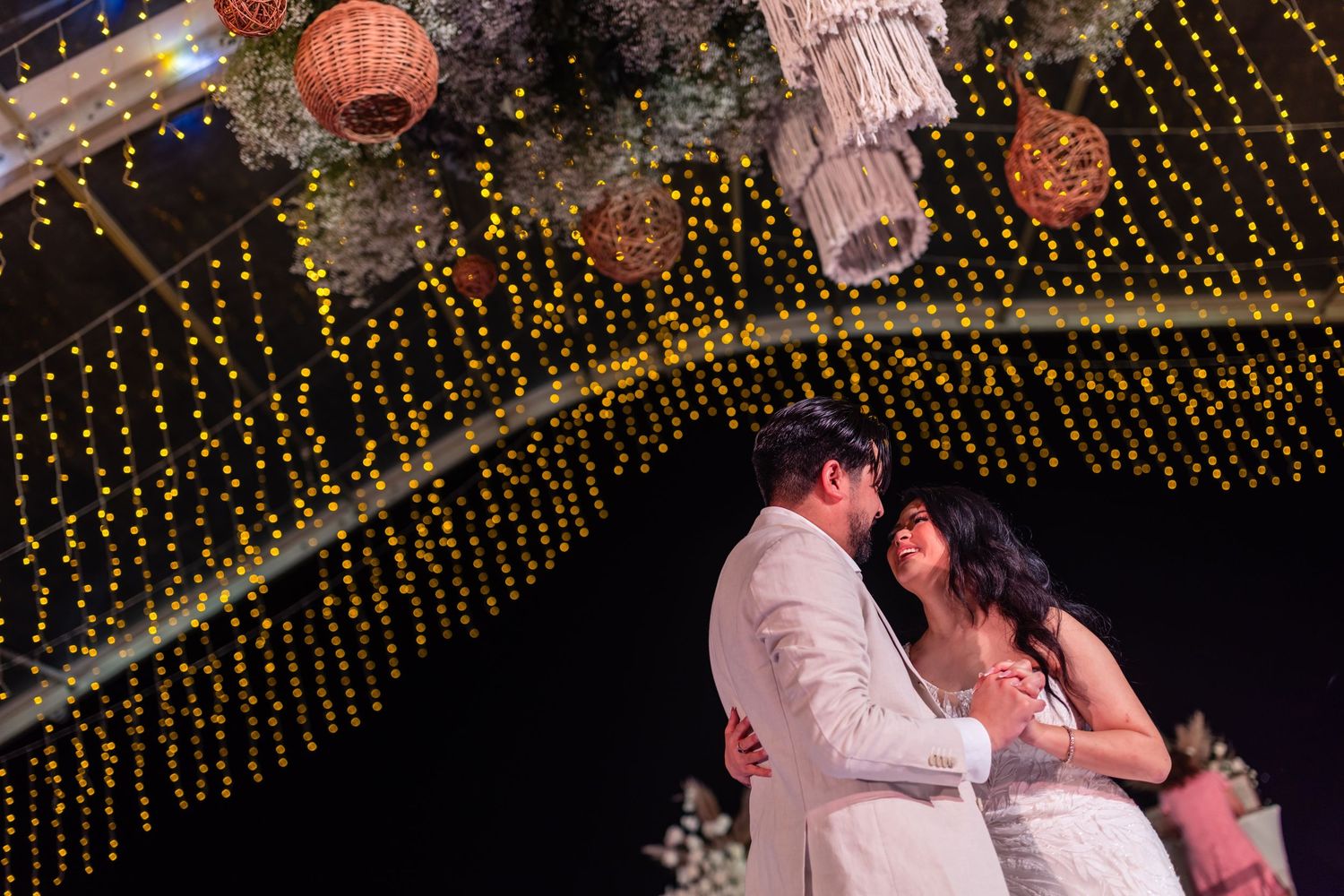 Romantic couple shares intimate dance under glowing fairy lights and floral decorations at evening wedding reception.
