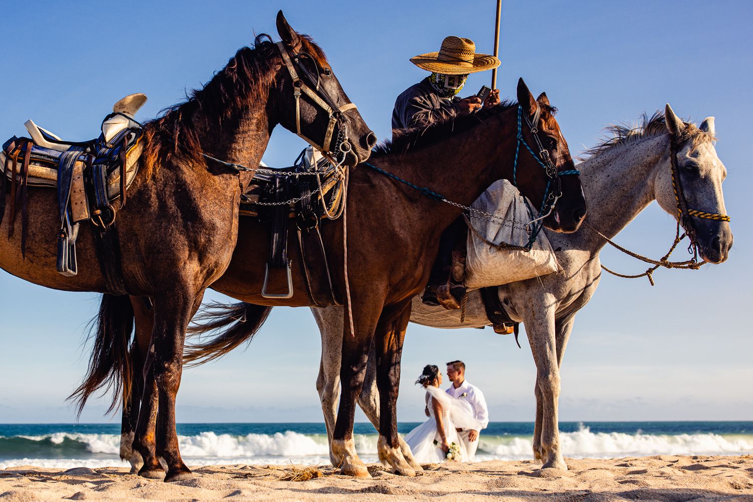 A cowboy on horseback with two saddled horses on a sunny beach, couple embracing in the background.