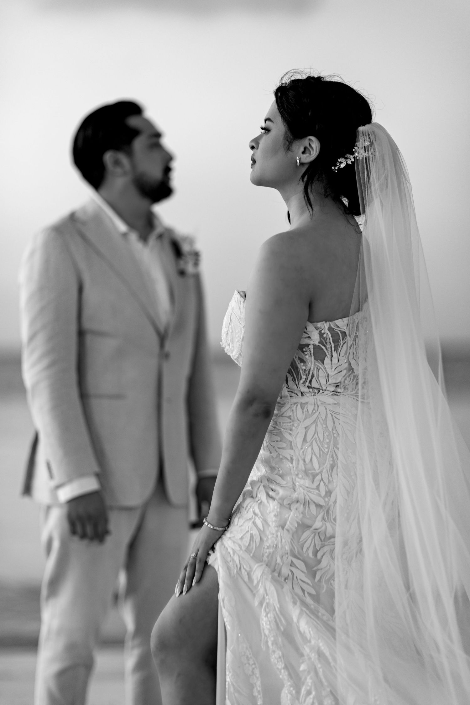 Black and white photo of bride in lace gown with veil gazing up while groom stands blurred in background.