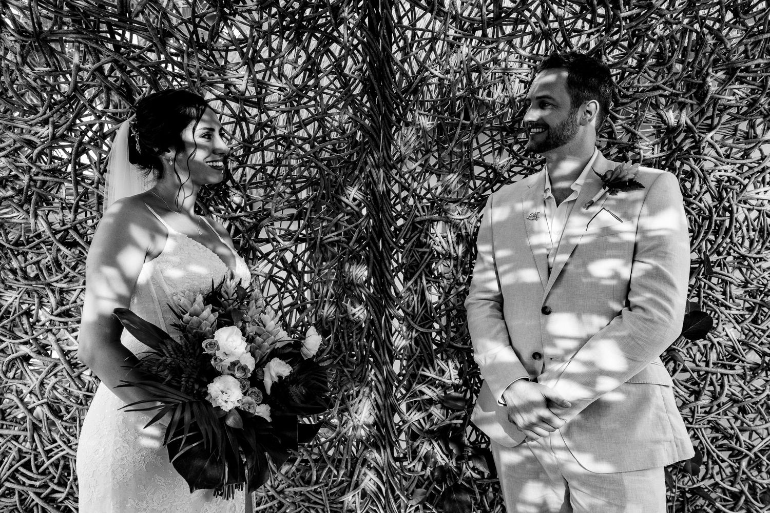 Black and white photo of a bride holding bouquet and groom in suit standing together at an outdoor wedding ceremony.
