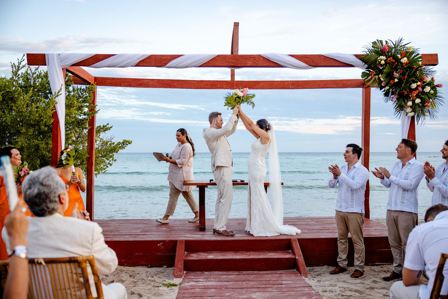 Couple raises bouquet at beach wedding ceremony under red canopy with ocean backdrop.