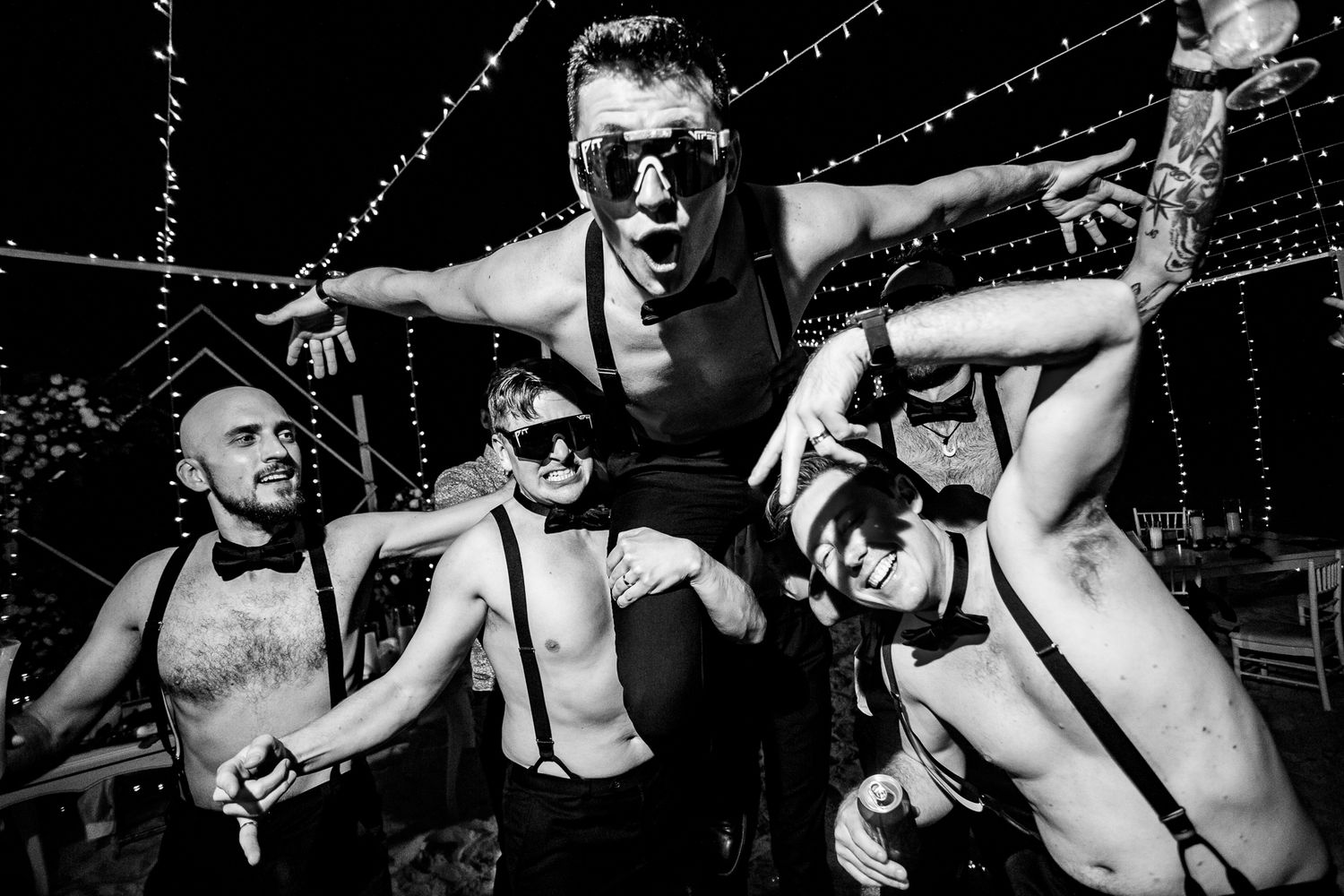 Black and white photo of four shirtless men posing energetically outdoors at night under string lights.