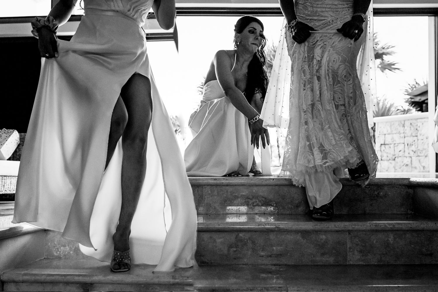 Black and white photo of bridesmaids in white gowns on steps, bride crouching in background at outdoor wedding.