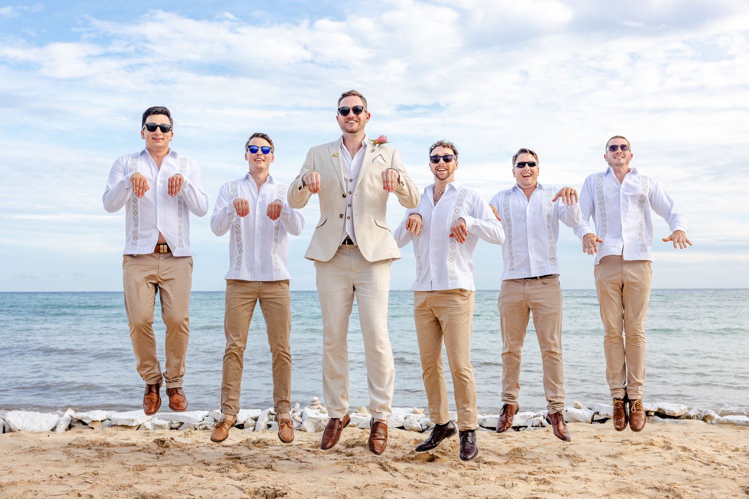 Groomsmen in white shirts and khaki pants jumping on a beach with the groom in a tan suit on a sunny day.
