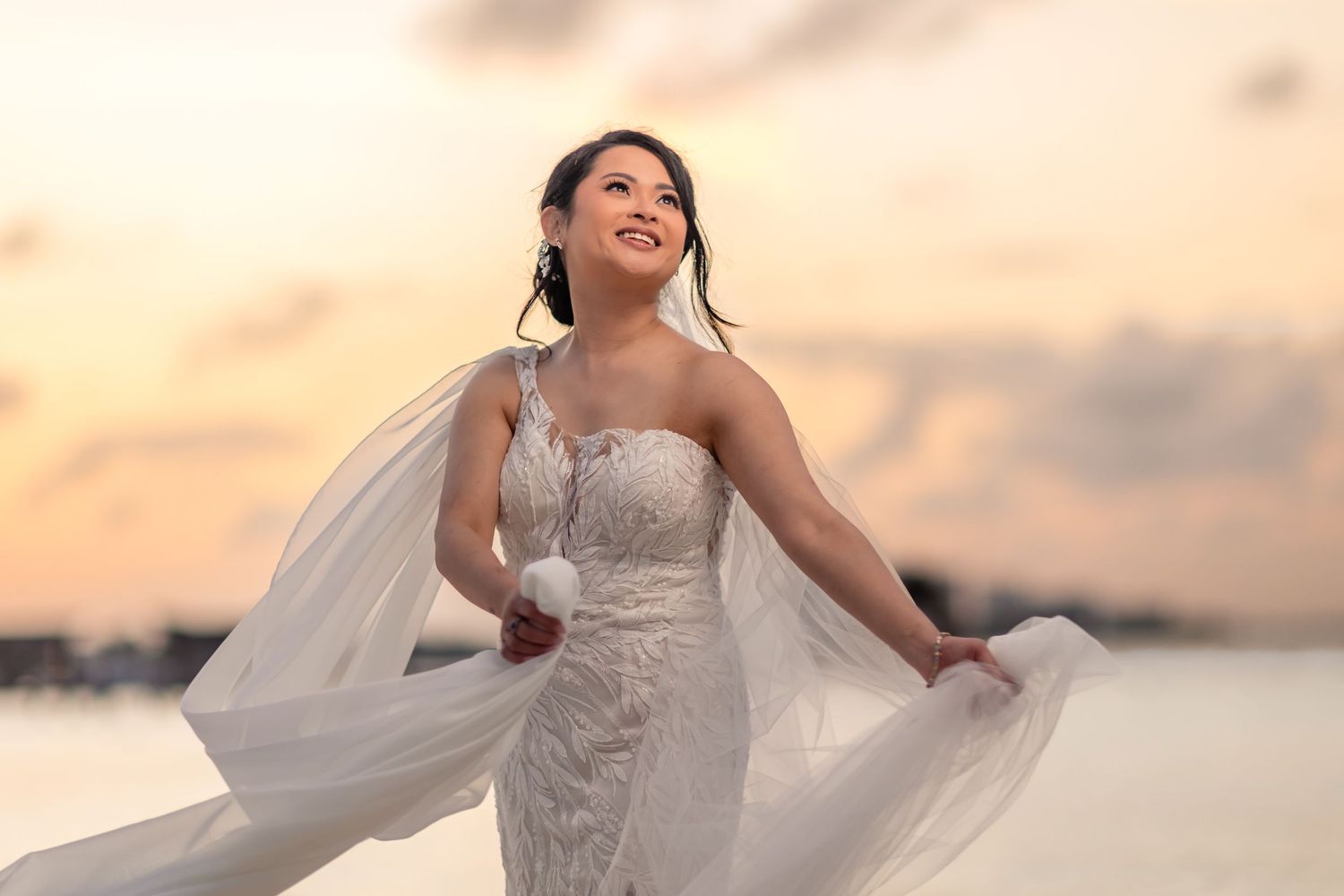 Bride in white lace gown with flowing veil poses at sunset beach, smiling joyfully.