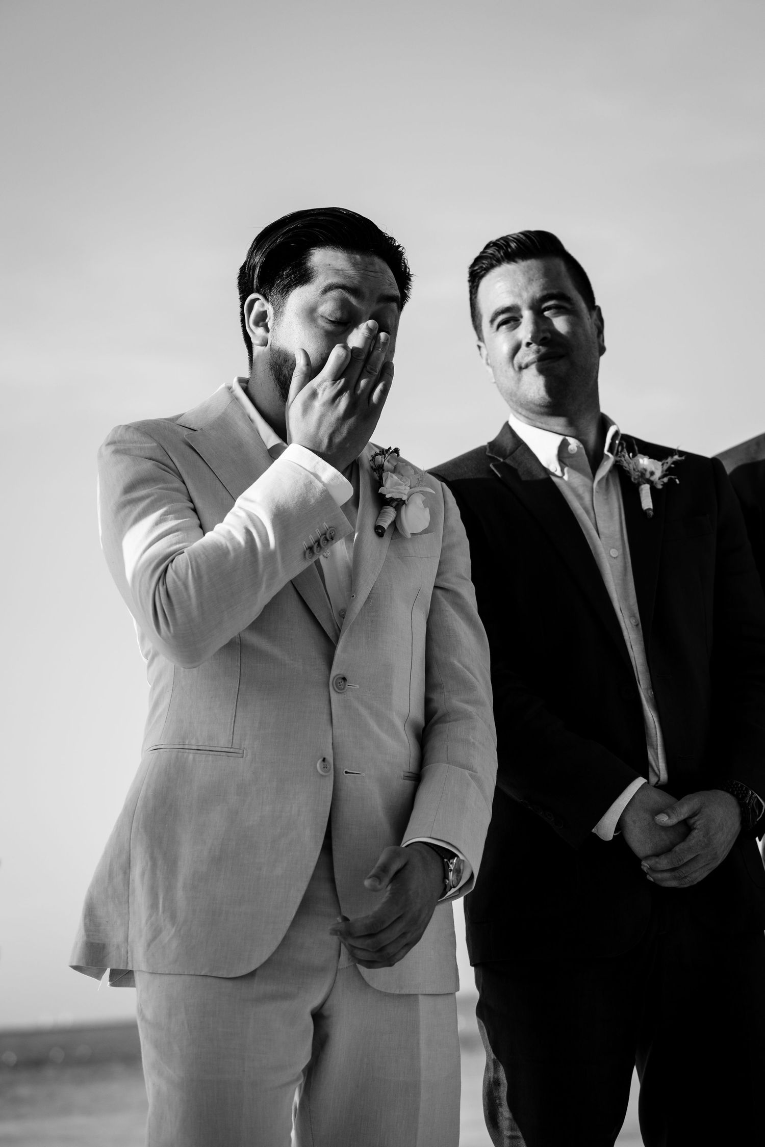 Emotional groom covering mouth in tears beside smiling groomsman in black and white wedding photo.