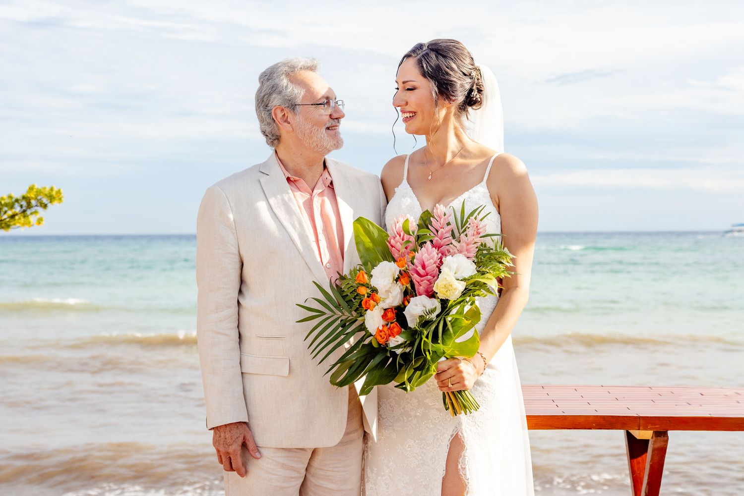 Bride and groom smiling at each other on a tropical beach, bride holding a vibrant bouquet of tropical flowers.