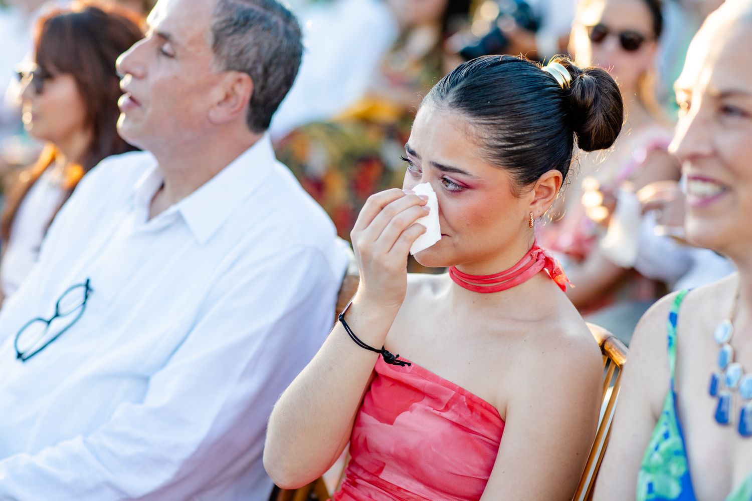 Emotional woman in red strapless dress wiping tears at outdoor event, with blurred guests seated nearby.