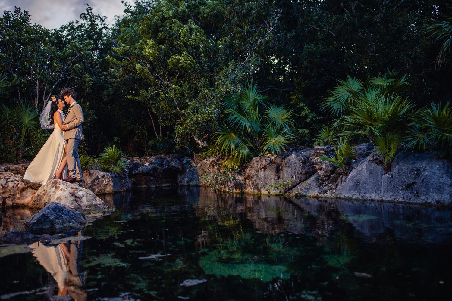 Bride and groom share a romantic kiss on rocks beside a serene jungle cenote, with lush tropical greenery reflected in the water.