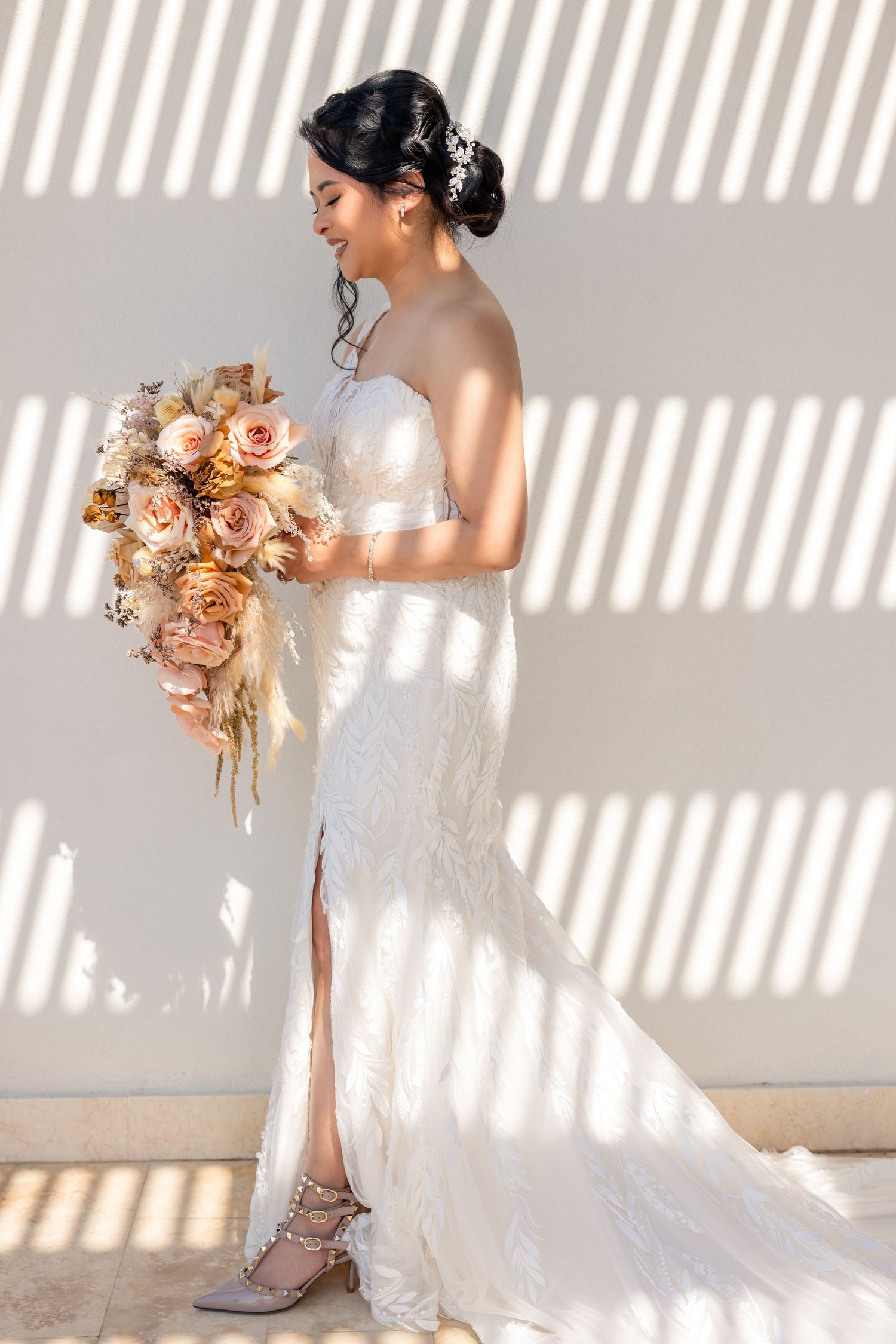 Elegant bride in strapless lace gown holding dried floral bouquet, standing in striped sunlight against white wall.