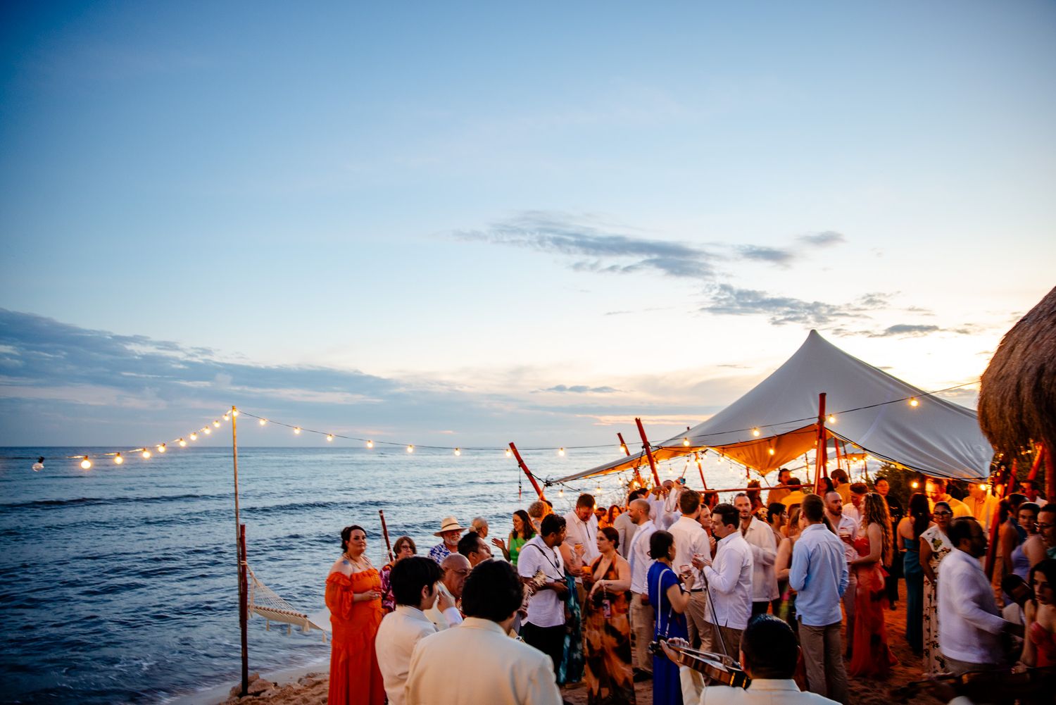 Beach wedding reception at sunset with string lights, tent, and guests gathered by the ocean.