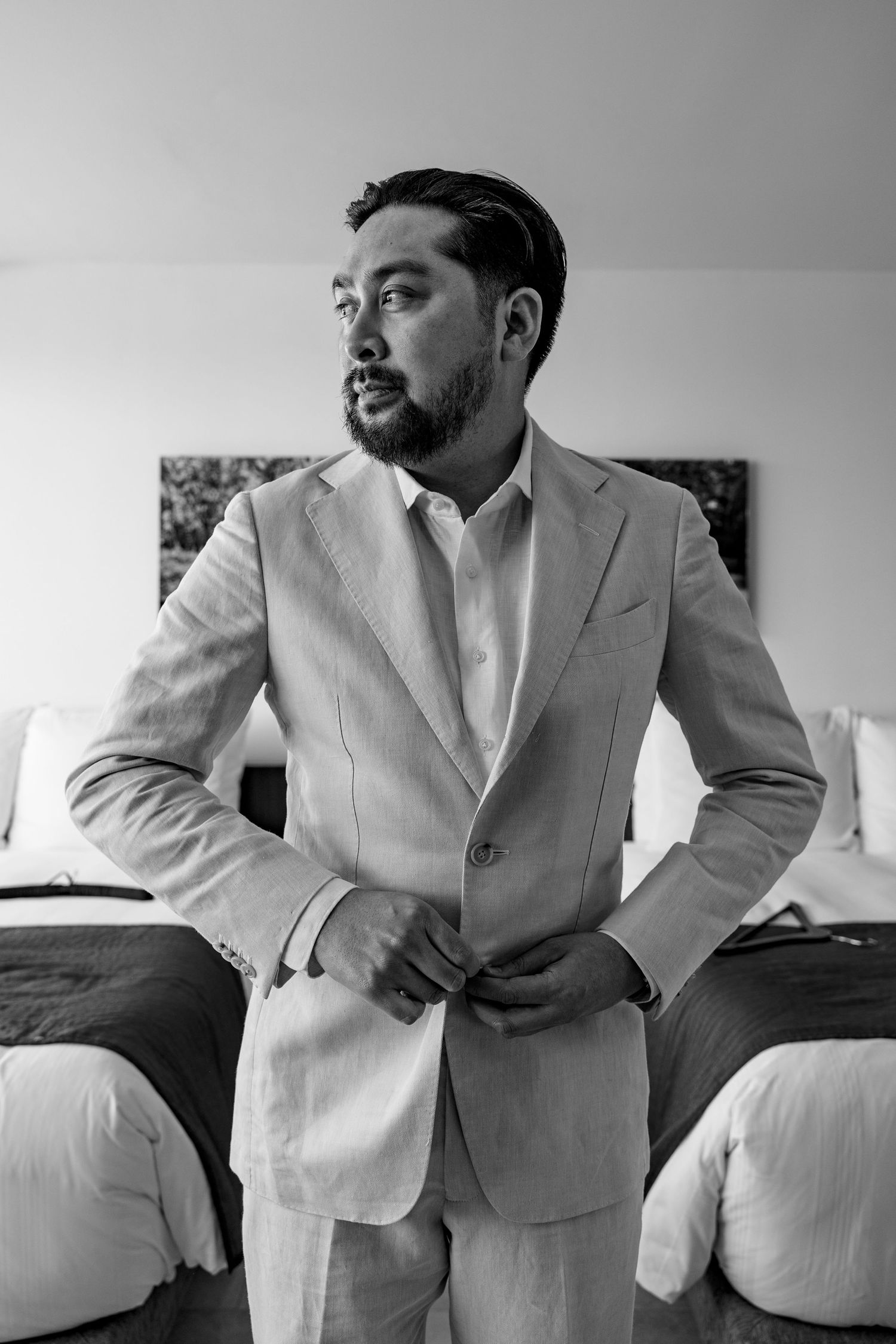 Black and white photo of a well-dressed man in a suit buttoning his jacket in a hotel room.