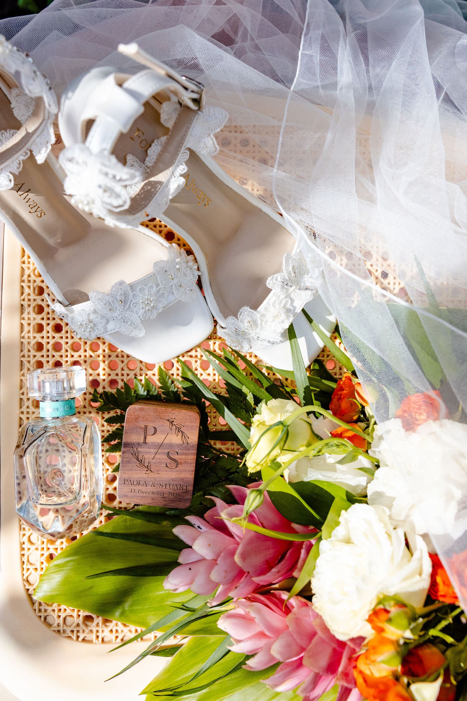 White lace bridal heels, perfume, wedding favor box, and tropical floral bouquet arranged on a rattan tray with veil.
