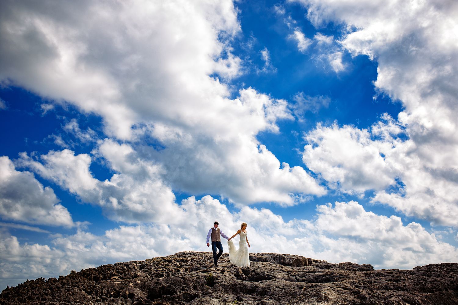 Bride and groom stand atop rocky cliffs under dramatic blue sky with white clouds on their wedding day.