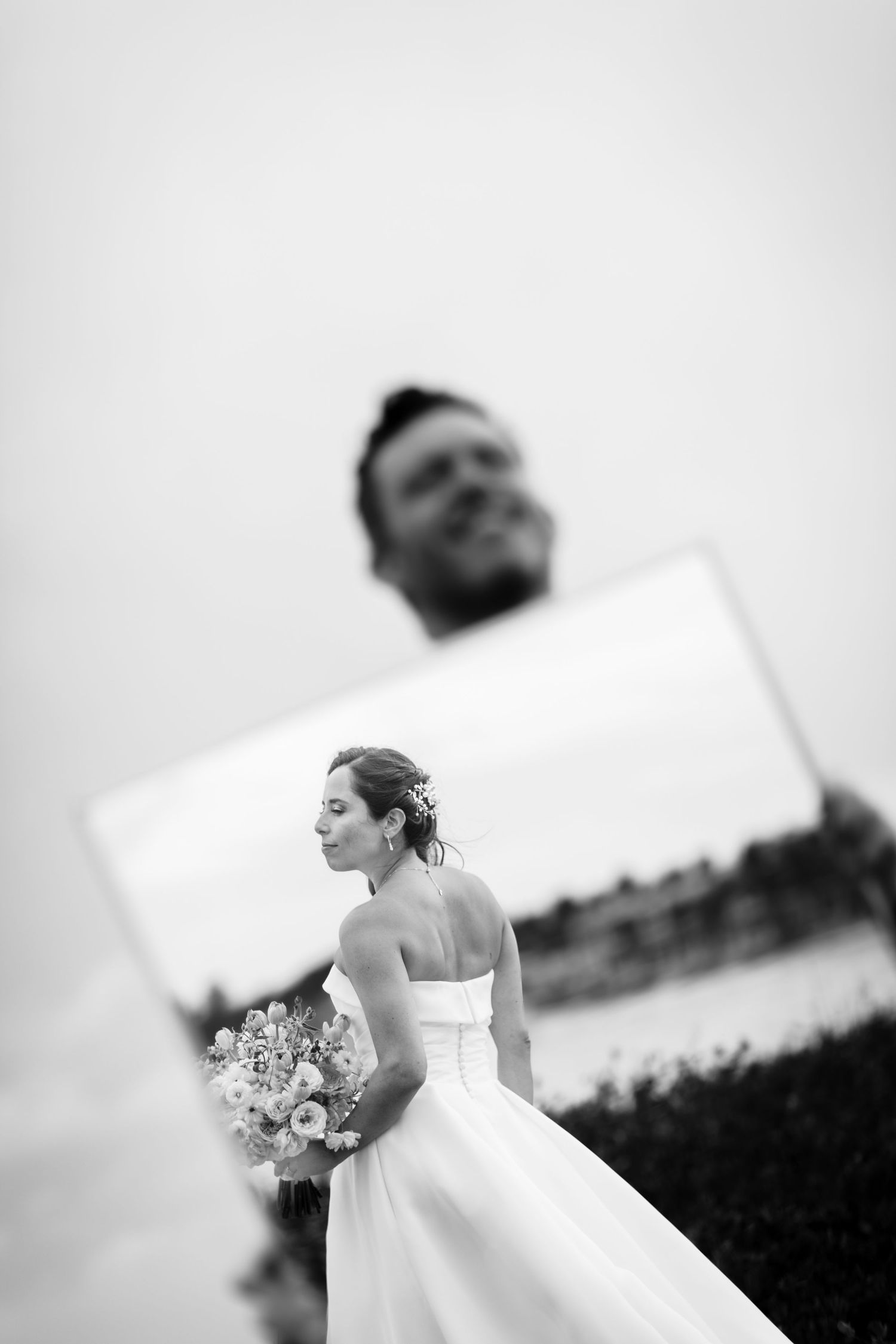 Black and white wedding photo of bride holding bouquet with groom's reflection in mirror, romantic outdoor portrait.