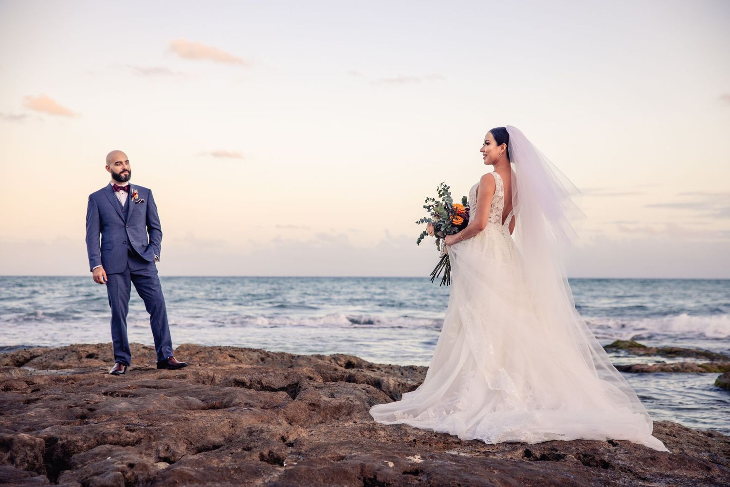 Bride in white gown with veil holds bouquet facing groom in blue suit on rocky beach at sunset.