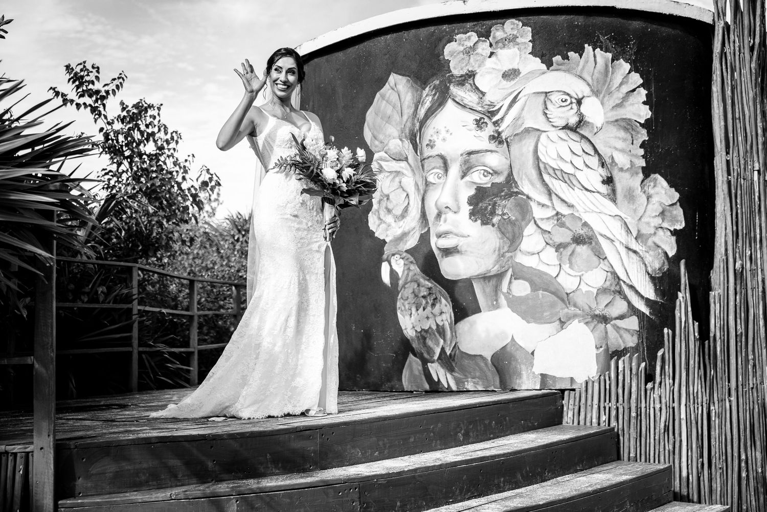 Bride in white gown waves beside a floral mural on steps, black and white wedding photo.