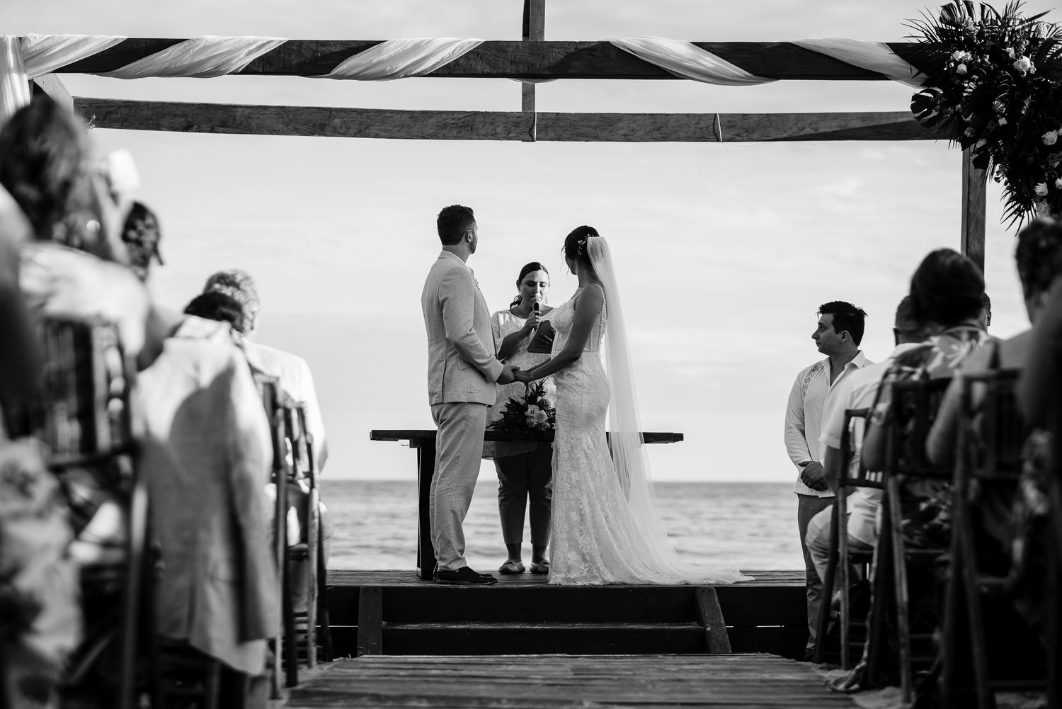 Black and white photo of a beach wedding ceremony with couple exchanging vows under a canopy by the ocean.