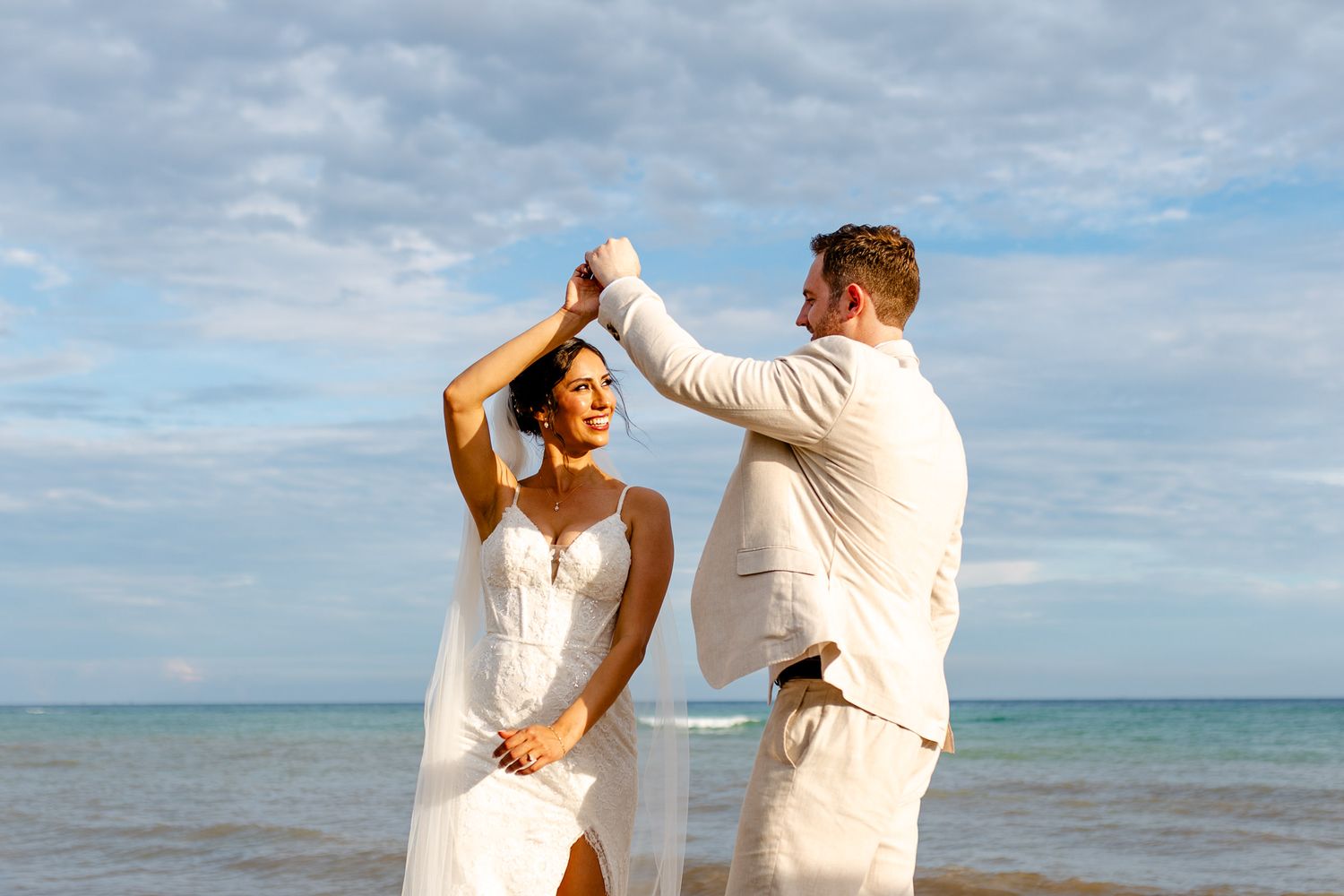 Bride and groom dancing on beach in white wedding attire with ocean backdrop.