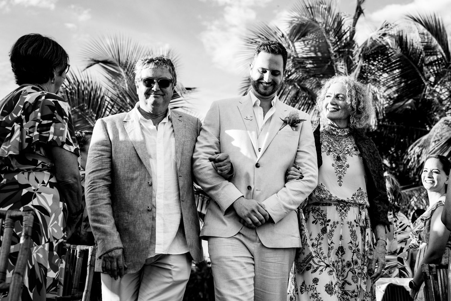 Black and white wedding photo of two grooms walking with an elderly woman amid tropical palms.