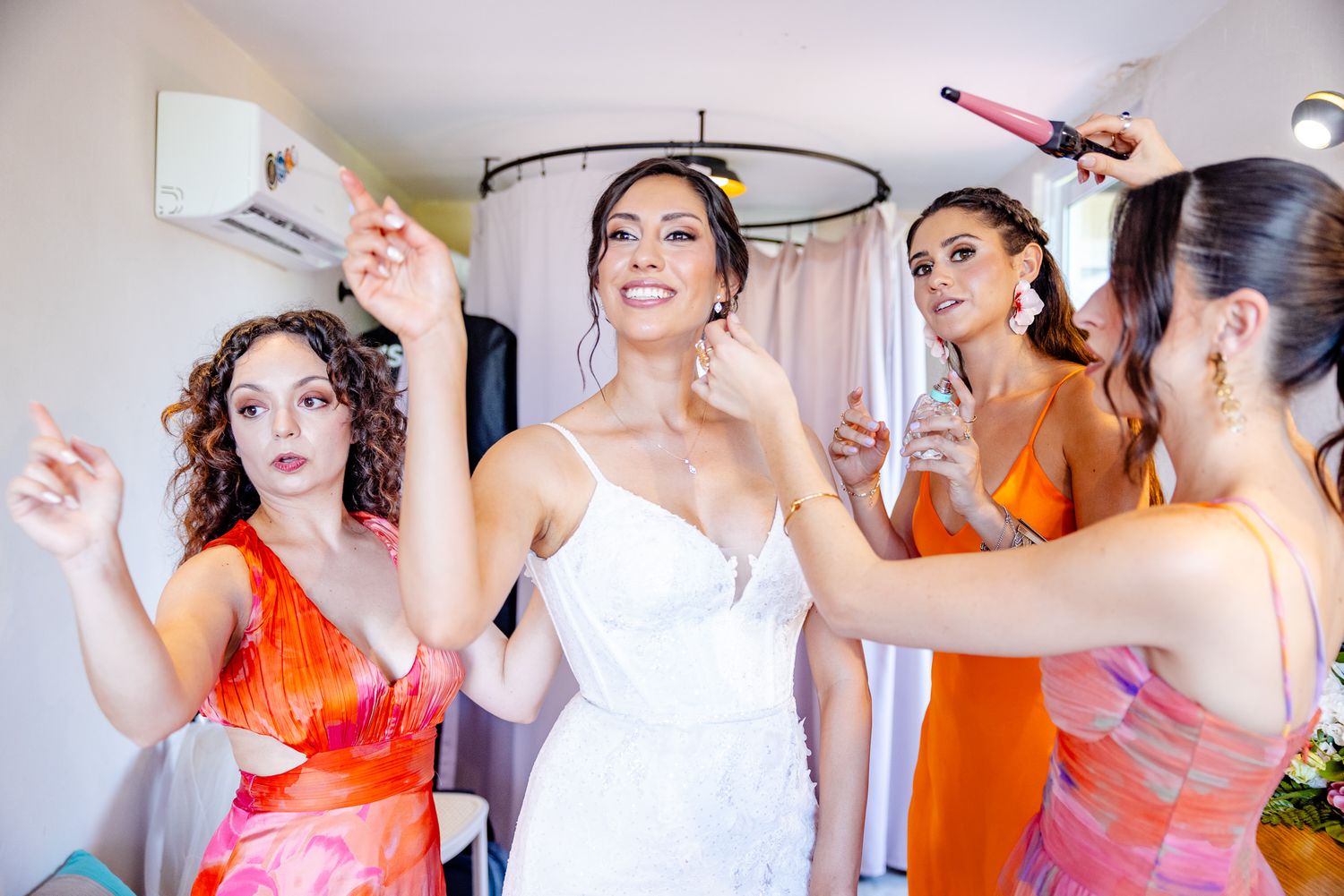 Bride in white gown surrounded by bridesmaids in orange dresses getting hair and makeup done before wedding.