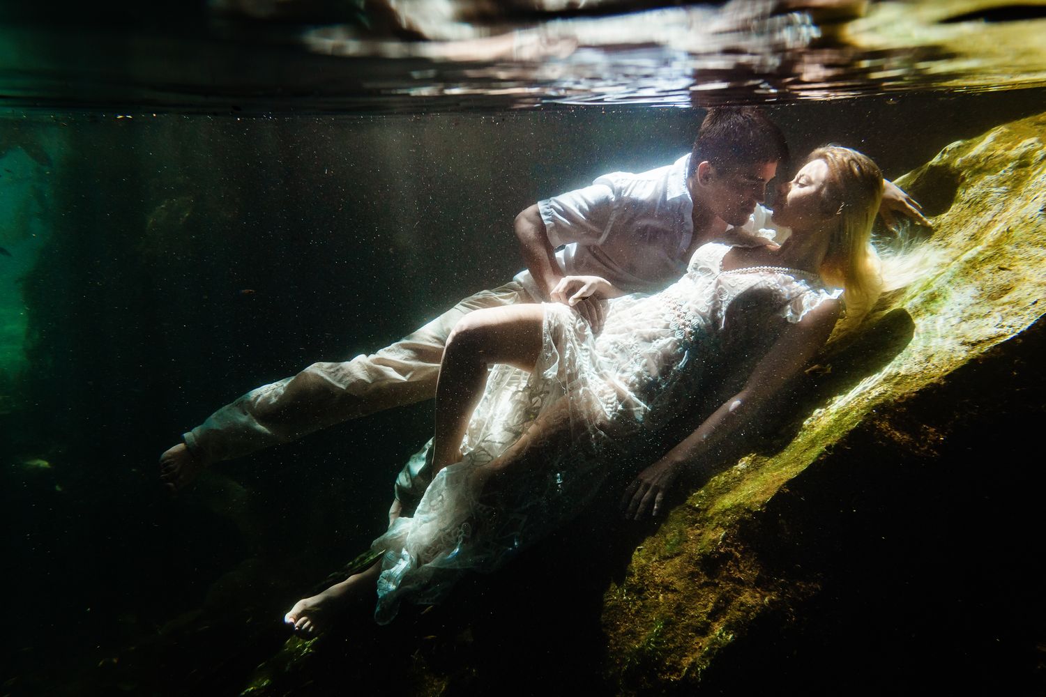 Couple in wedding attire posing romantically underwater in a cenote, surrounded by rocks and ethereal light.