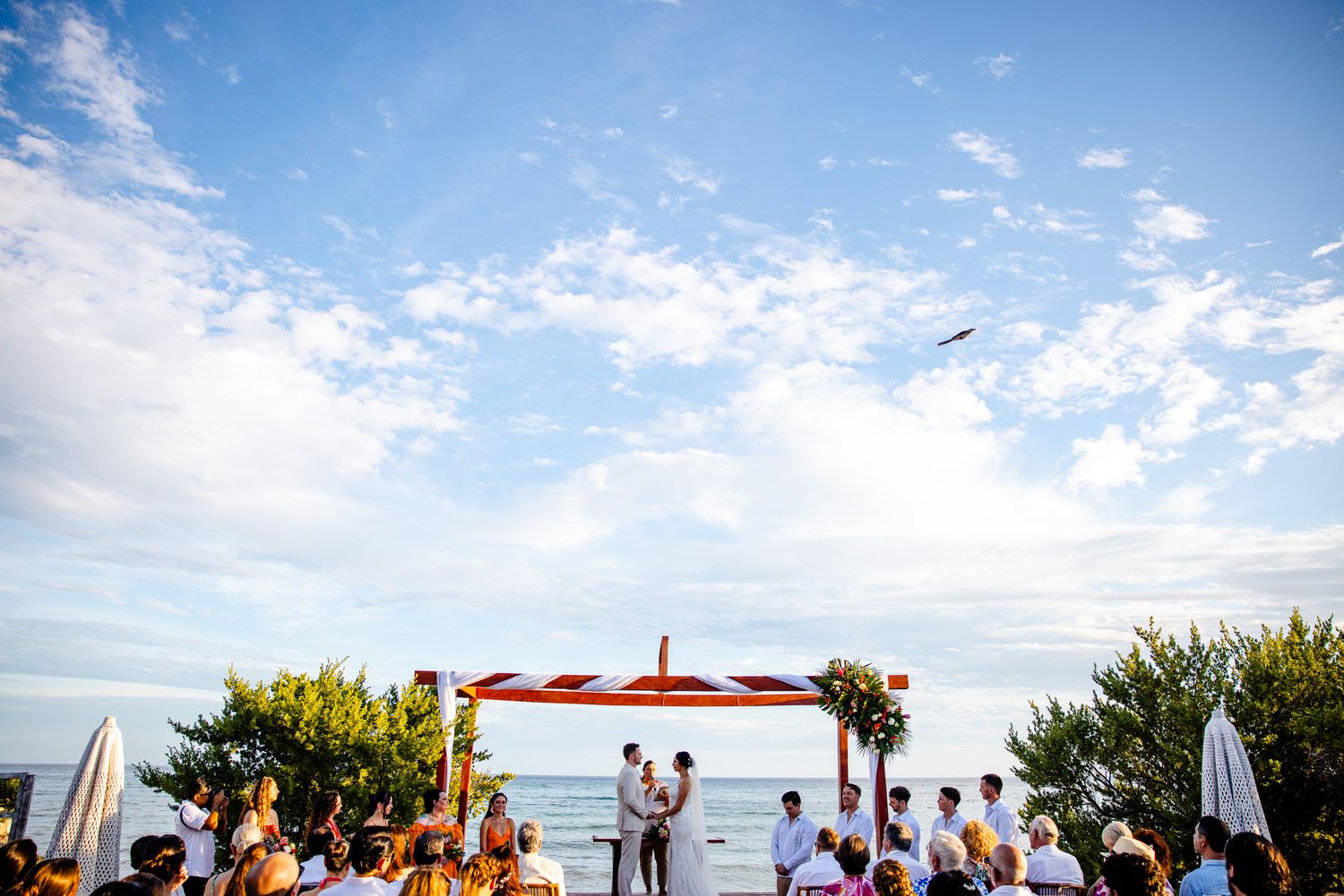 Outdoor beach wedding ceremony under a colorful canopy arch with guests seated by the ocean on a sunny day.