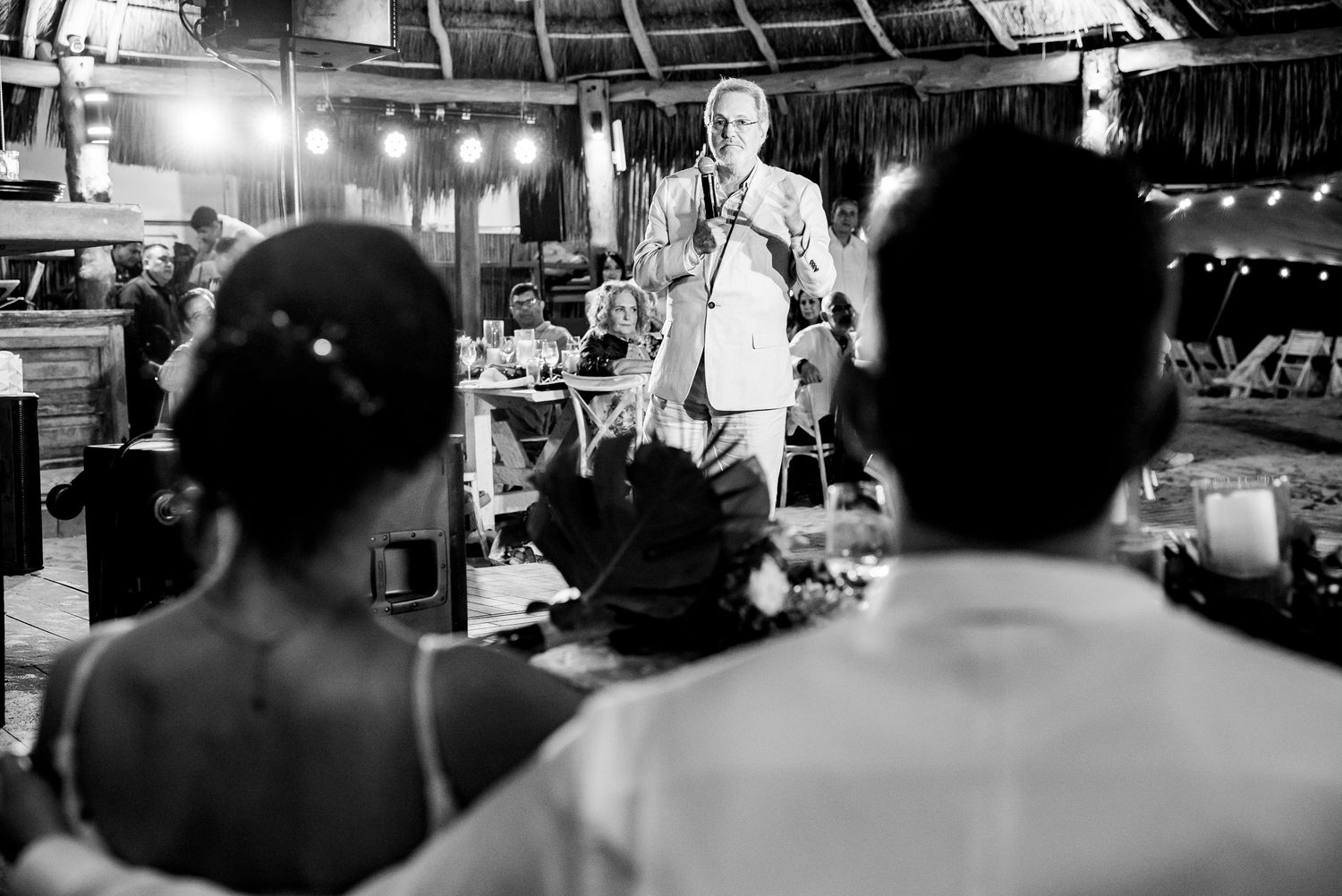 Black and white wedding photo of couple listening to an elder giving a heartfelt toast at a tropical outdoor reception.