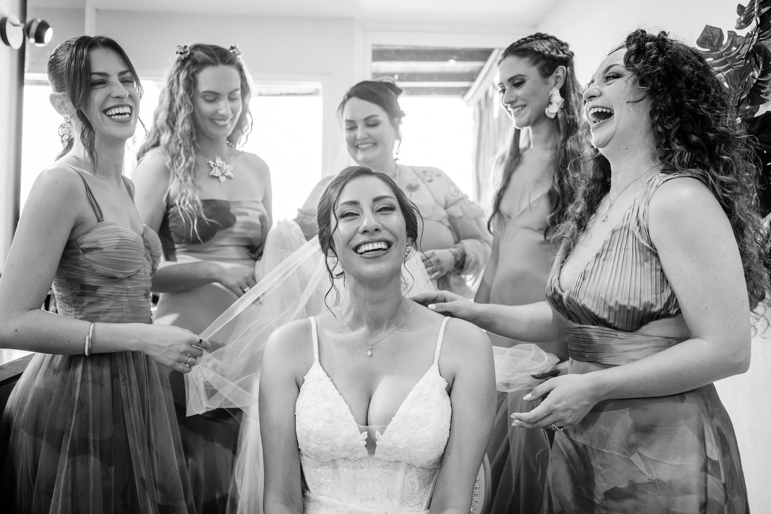 Bride laughing joyfully surrounded by smiling bridesmaids during wedding preparations in black and white photo.