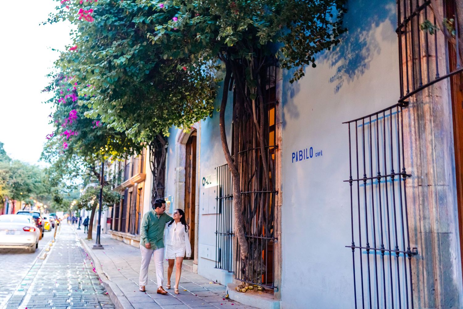 Couple strolling on a charming cobblestone street lined with trees and colorful buildings in a Latin American town.