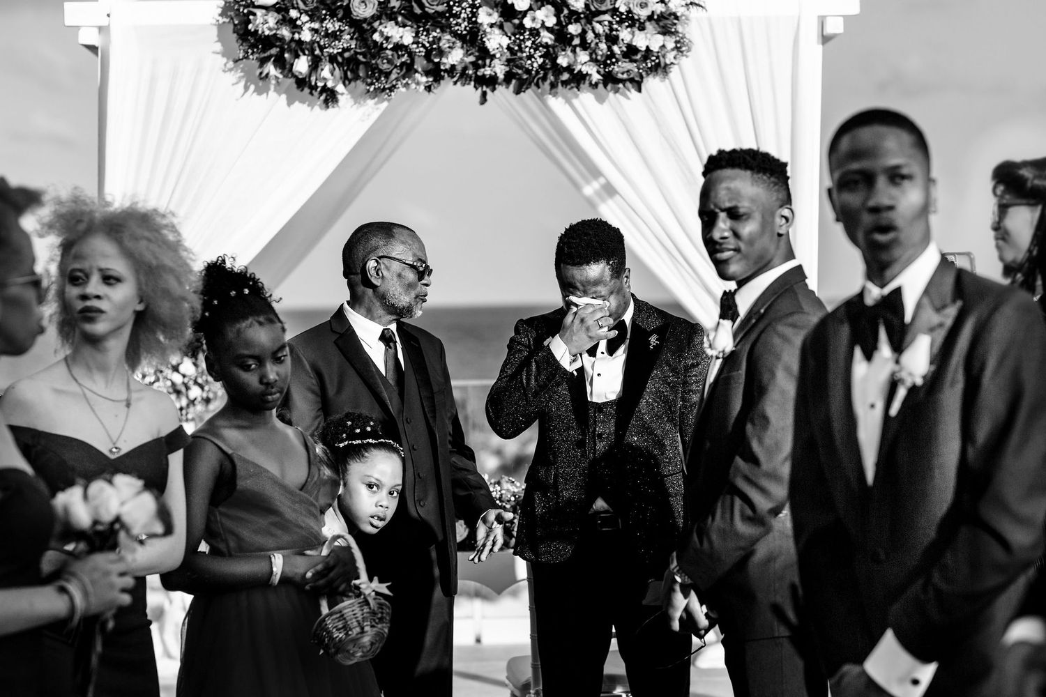 Black and white wedding ceremony photo showing groomsmen and guests gathered under a floral arch outdoors.