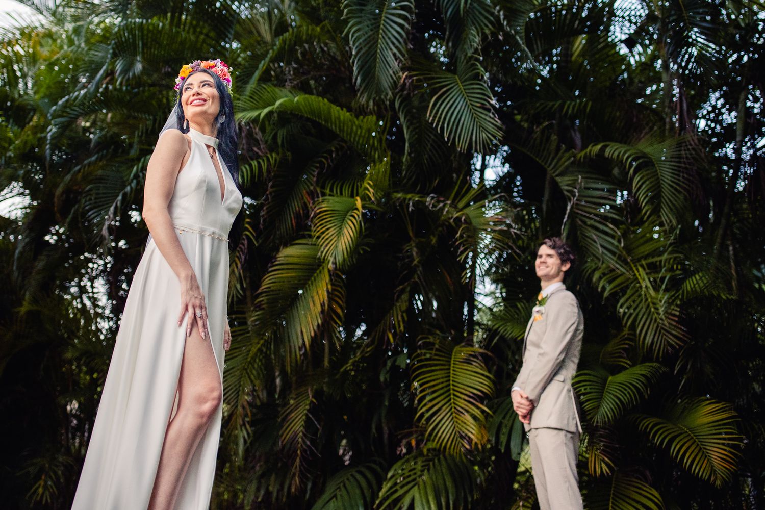 Bride in white gown with floral crown and groom in beige suit stand among lush tropical palm trees outdoors.