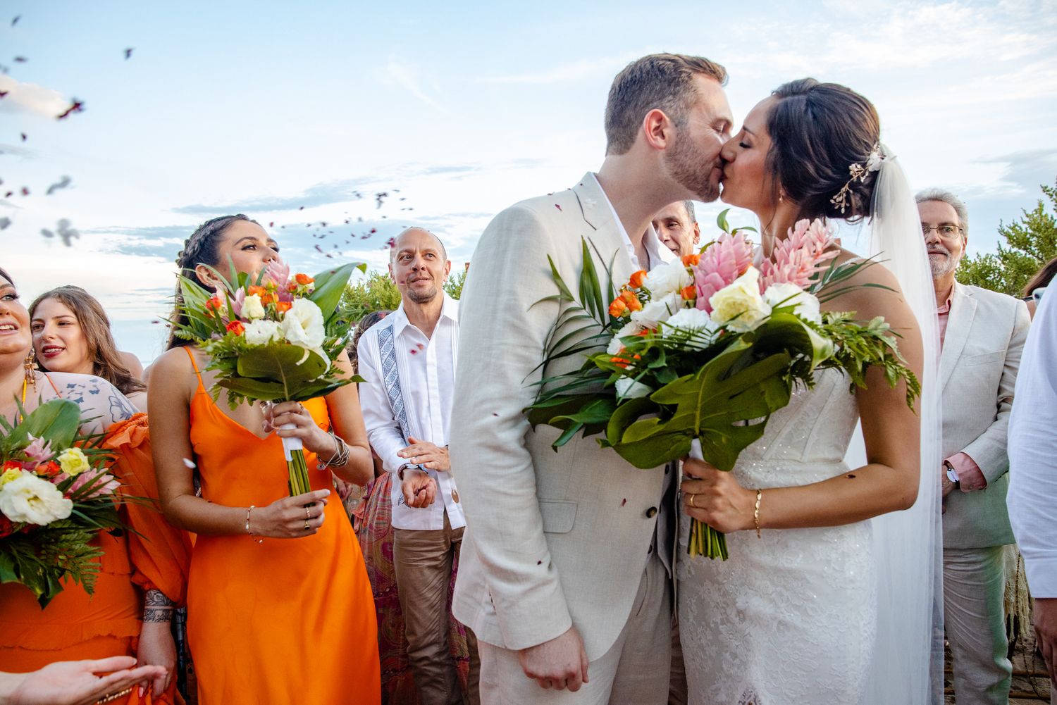 Bride and groom share first kiss at outdoor beach wedding ceremony with bridesmaids in orange dresses holding bouquets.