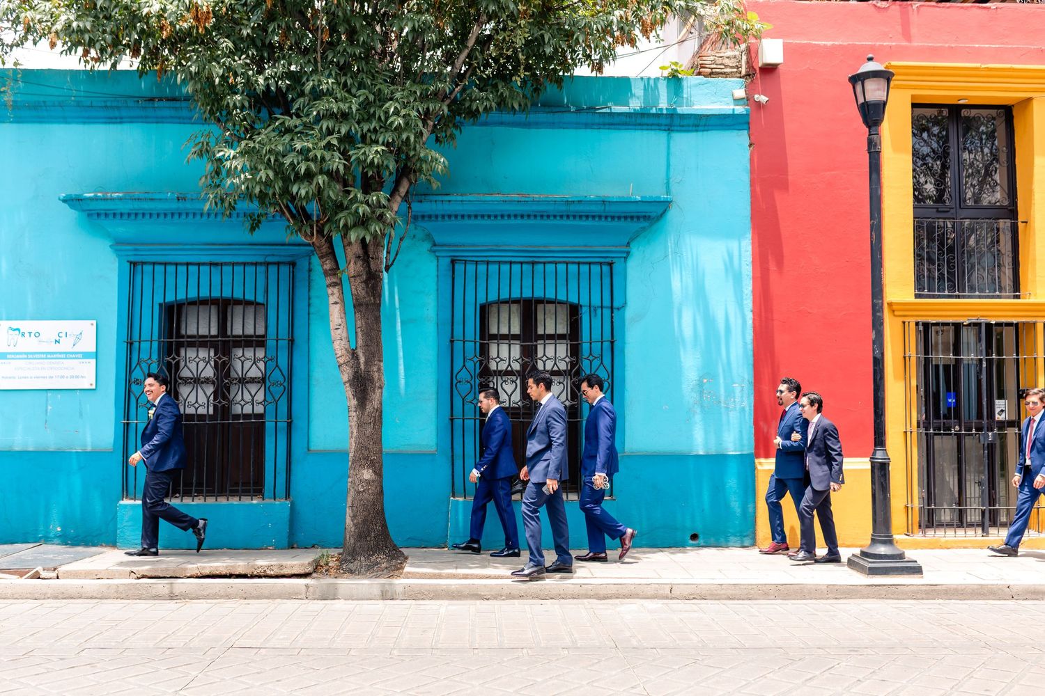 Men in blue suits walking past vibrant colorful buildings on a sunny street in Latin America.