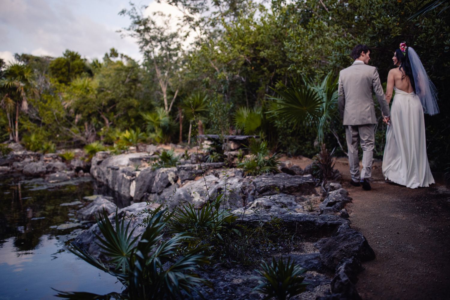Wedding couple walks hand-in-hand along rocky garden path surrounded by lush tropical greenery.