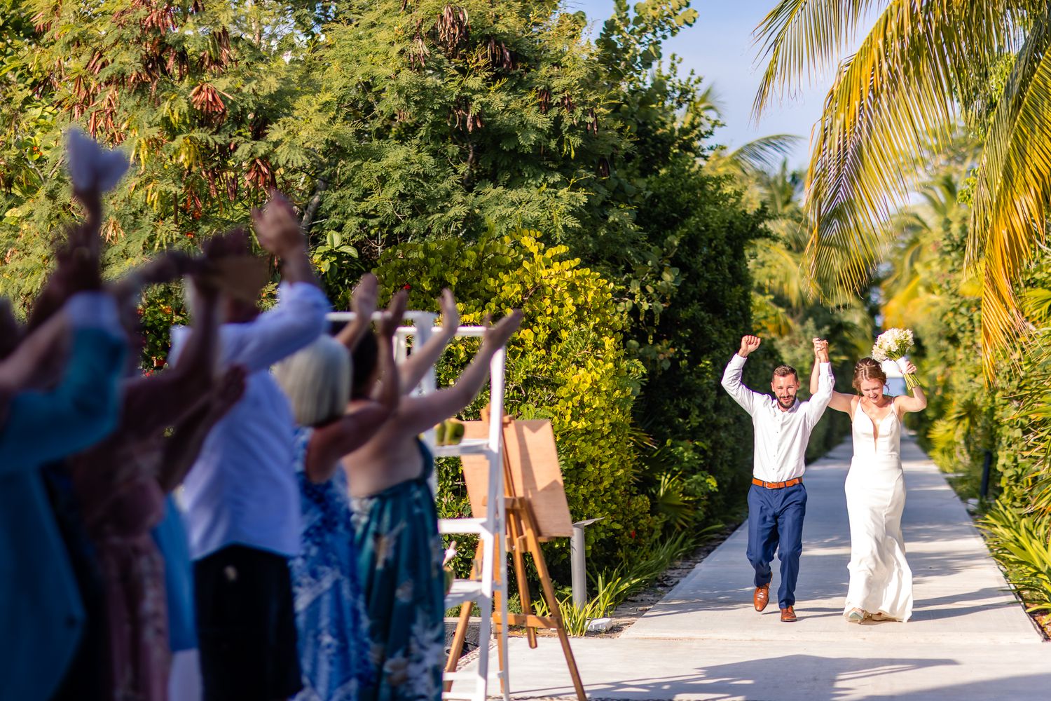 Joyful newlyweds walk down tropical path as guests celebrate their wedding ceremony outdoors.