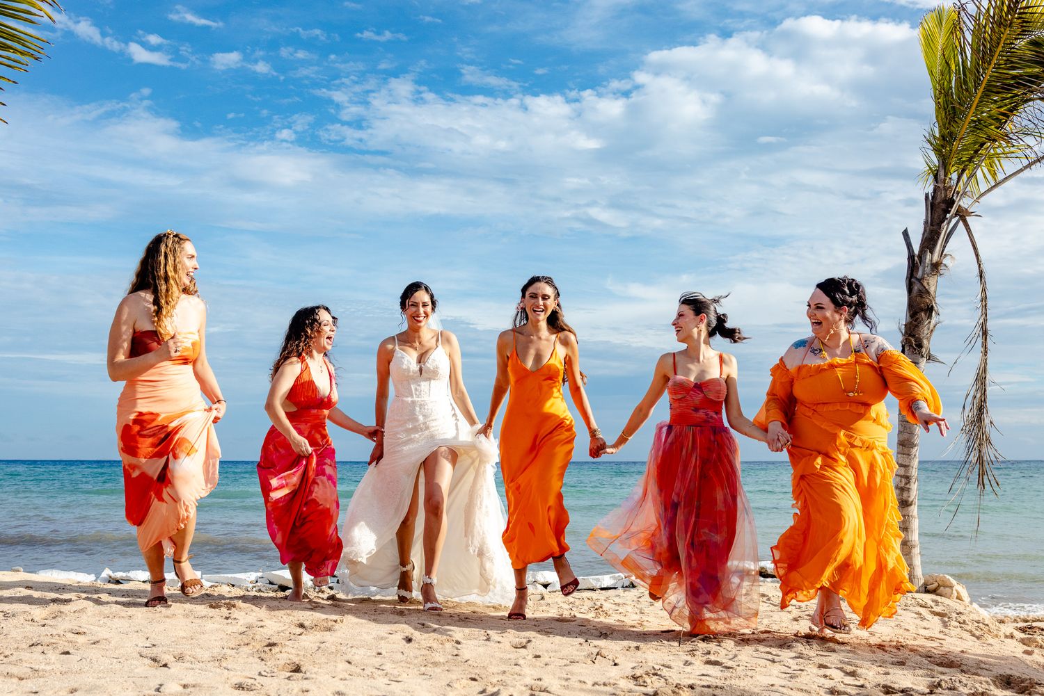 Bridesmaids in colorful orange and red dresses walk hand-in-hand with bride on tropical beach.