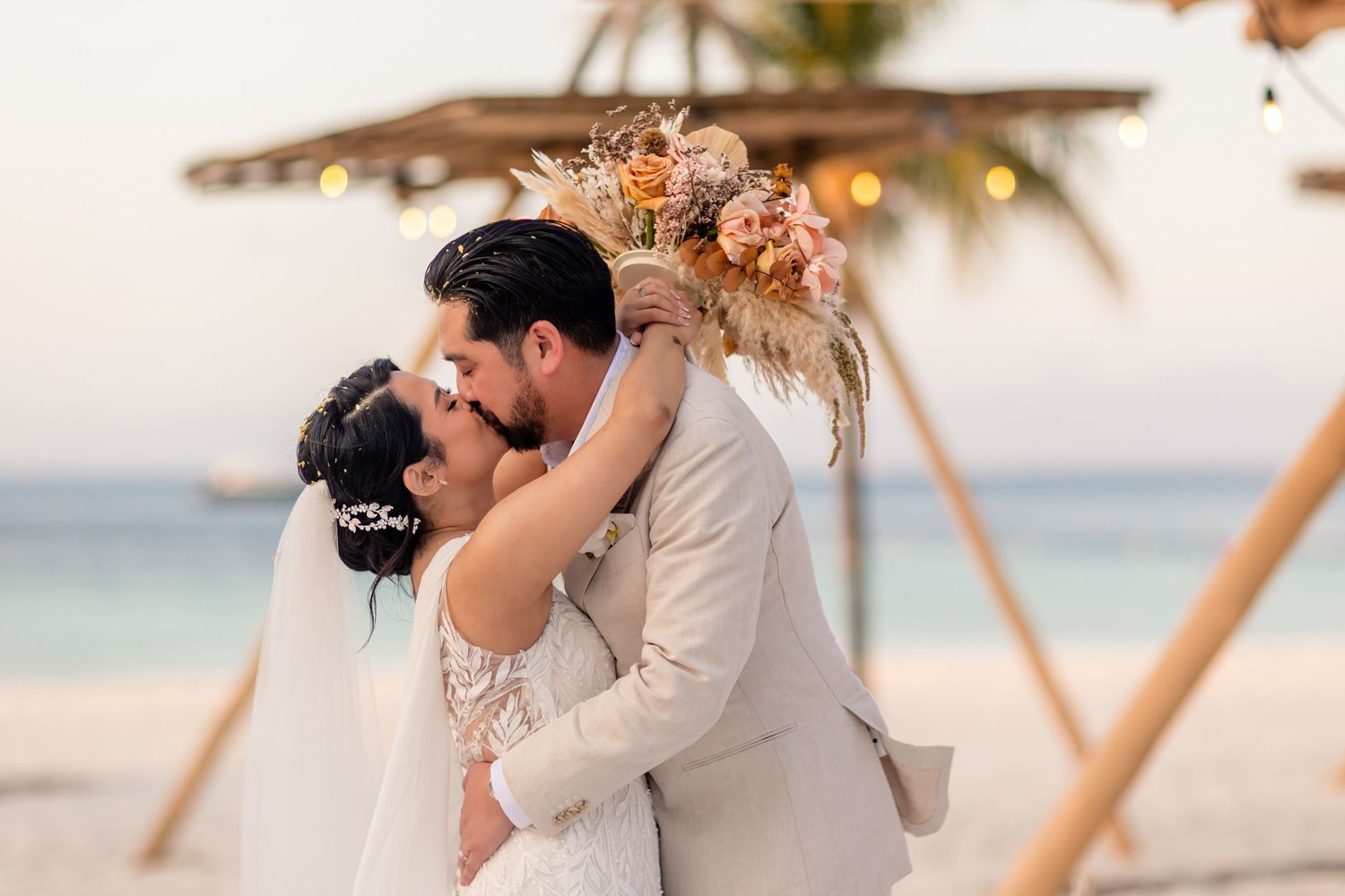 Bride and groom share first kiss at romantic beach wedding ceremony with boho floral arch and string lights.