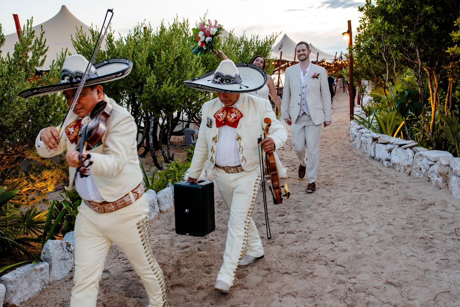 Mariachi band in traditional white charro suits walking along a sandy path at a Mexican outdoor event.